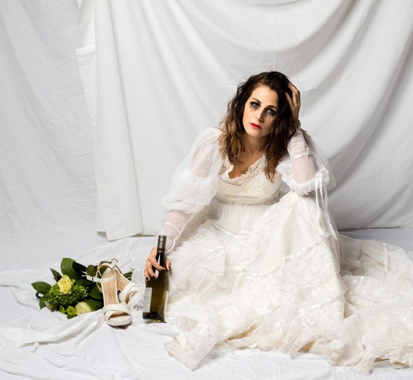 young woman with tear tracks sitting on ground wearing an 80s wedding dress with flowers shoes off and bottle of wine