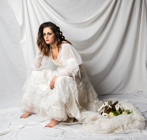 young woman with mascara tear tracks sitting on ground wearing an 80s wedding dress with flowers shoes off and bottle of wine